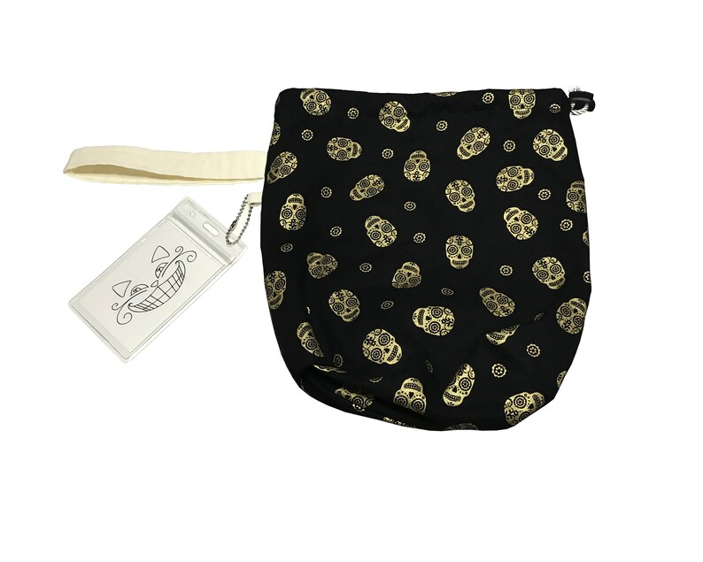 May include: Black drawstring bag with a gold sugar skull pattern. The bag has a white strap and a clear plastic tag with a black and white illustration of a smiling face.