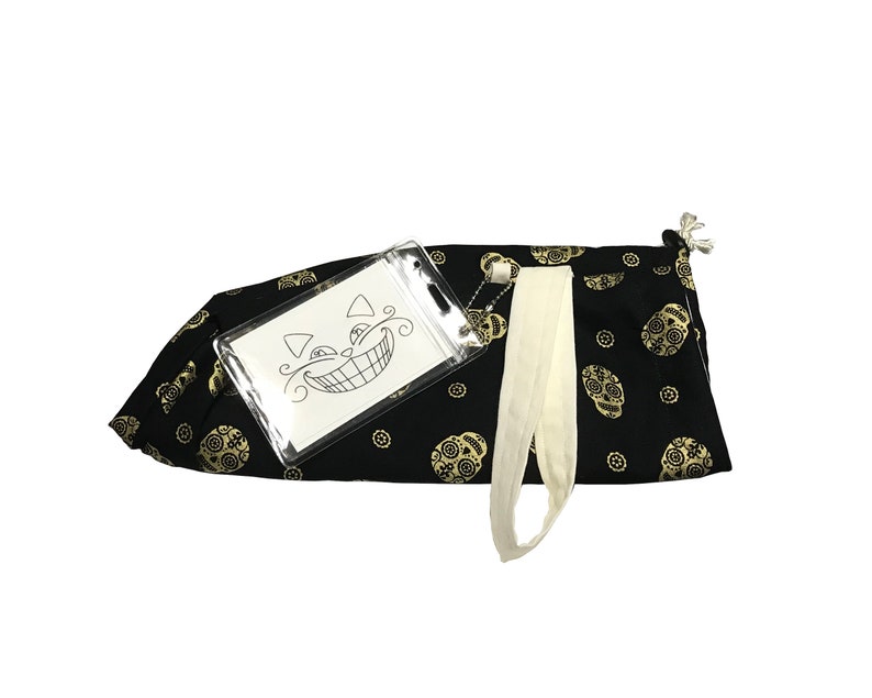 May include: Black drawstring bag with a gold skull pattern. A clear plastic pouch with a black and white illustration of a cat with a wide grin is attached to the bag.