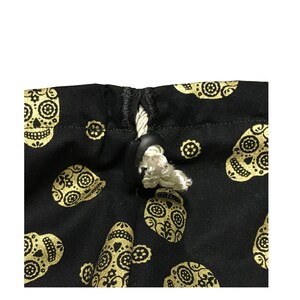 May include: A black drawstring bag with a white cord and a black toggle closure. The bag is decorated with a repeating pattern of gold sugar skulls.