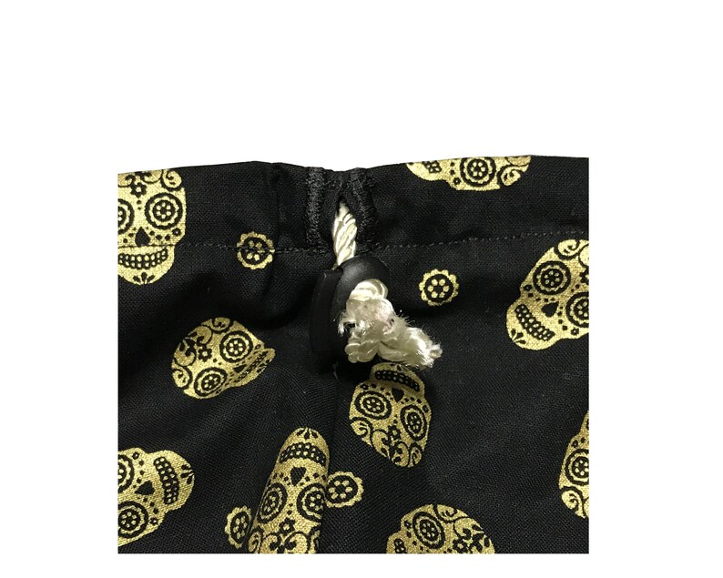 May include: A black drawstring bag with a white cord and a black toggle closure. The bag is decorated with a repeating pattern of gold sugar skulls.