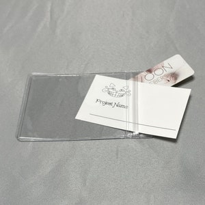 May include: A clear plastic card holder with a white card inside. The card has the text "Project Name" and a smiling cat logo. A pink and white card with the text "LOON FIBERS" is partially visible behind the white card.