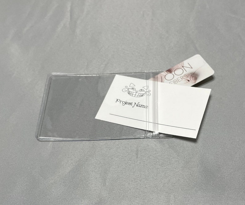 May include: A clear plastic card holder with a white card inside. The card has the text "Project Name" and a smiling cat logo. A pink and white card with the text "LOON FIBERS" is partially visible behind the white card.