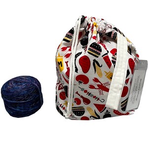 May include: A white drawstring bag with a red, yellow, and black print of various food items, including a burger, corn, and a lobster. The bag is sitting next to a ball of blue yarn.