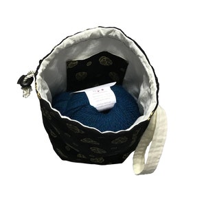 May include: A black drawstring bag with a gold floral pattern and a white cotton strap. The bag is open and a teal ball of yarn is inside.