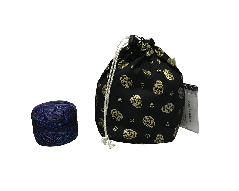 May include: A black drawstring bag with a gold skull pattern and a white drawstring. The bag is filled with a skein of blue and purple yarn.