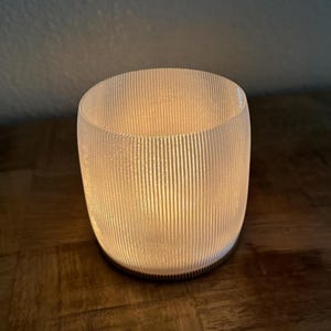 Op de afbeelding: Een witte, geribbelde glazen kaarsenhouder met een warme gloed die van binnenuit straalt. De houder staat op een houten oppervlak.