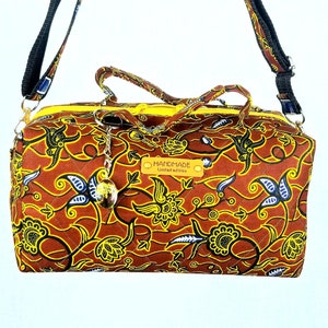 Könnte beinhalten: Eine braune gesteppte Handtasche mit gelbem Futter und einem verstellbaren schwarzen Trageriemen. Die Tasche hat ein Blumenmuster mit blauen und gelben Akzenten. Die Tasche hat ein Etikett mit der Aufschrift "HANDMADE limitierte Auflage".