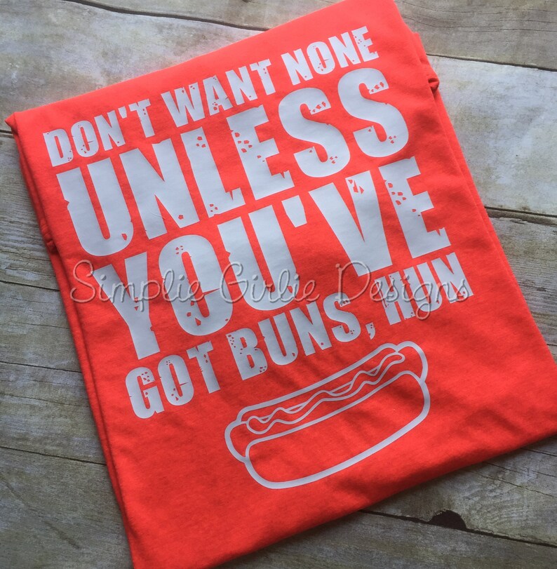 May include: A vibrant orange t-shirt featuring the white text "DON'T WANT NONE UNLESS YOU'VE GOT BUNS, HUN" and a hot dog graphic. The shirt is folded on a wooden surface.