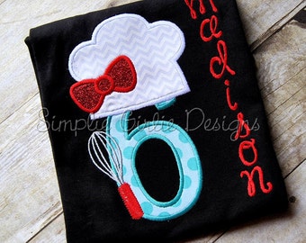 May include: Black t-shirt with a turquoise number six with white polka dots, a red whisk, and a white chef's hat with a gray chevron pattern and a red bow. The name "Madison" is embroidered in red on the side of the shirt.