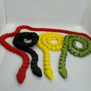 May include: Four brightly coloured, segmented snake toys in red, black, yellow, and green. Each snake is coiled and has a distinct head shape. The toys are arranged on a white surface, showcasing their vibrant colours and unique design.