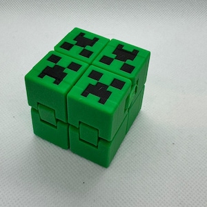 May include: A green fidget cube with black Creeper faces on each side. The cube is made of plastic and has a textured surface.