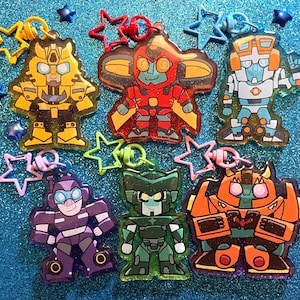 May include: Seven colorful keychains featuring different Transformers characters. The characters are Optimus Prime, Bumblebee, Megatron, Starscream, Soundwave, Ironhide, and Ratchet. Each keychain is made of clear plastic with a metallic finish.