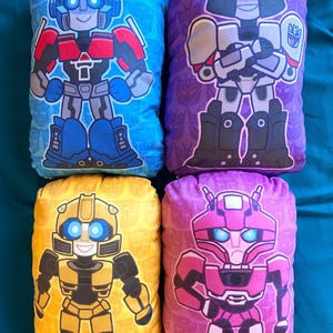 May include: Four plush pillows featuring characters from the Transformers franchise. The pillows are rectangular and feature a blue, purple, yellow, and pink background. Each pillow features a different character: Optimus Prime, Megatron, Bumblebee, and Arcee.