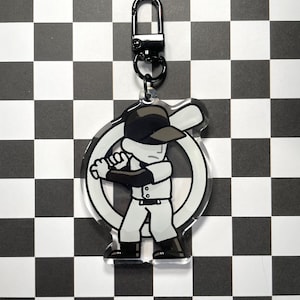 May include: A baseball-themed keyring with a cartoon batter in a white uniform and a black cap, set against a white circle. The keyring has a silver clasp and a black ring. The background is a black and white checkered pattern.