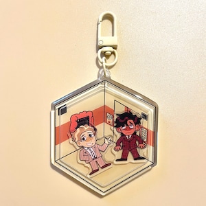 May include: A clear, hexagonal keychain with a cartoon elevator scene. Two figures are depicted, one pointing, the other looking worried. The keychain has a silver border and a cream-coloured clasp.
