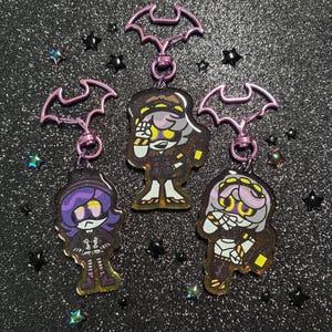 Peut inclure: Trois porte-clés en acrylique représentant des personnages de dessin animé stylisés avec des cheveux violets, des yeux jaunes brillants et des tenues sombres. Chaque breloque est fixée à un fermoir métallique violet en forme de chauve-souris.