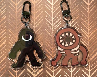 May include: Two acrylic keychains featuring cartoon monster designs. One is a dark green figure with a single white eye, and the other is a brown creature with a large mouth and teeth. Both have black metal clasps.