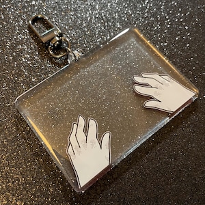 May include: A clear, rectangular keychain with a silver glitter background and a silver clasp. The keychain features two white hand illustrations with black outlines. The hands are reaching towards each other.