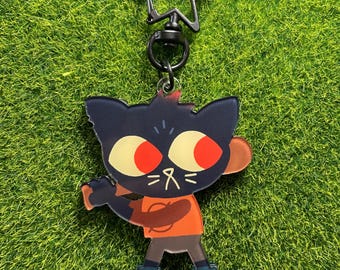 May include: A black acrylic keychain featuring a cartoon cat character with large red eyes, wearing an orange shirt and blue shoes. The keychain has a star-shaped clasp.