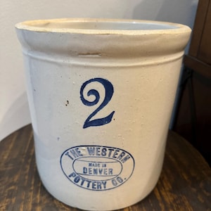 May include: A white ceramic crock with blue lettering. The crock is labeled "The Western Pottery Co. Made in Denver" and has the number "2" on the front.