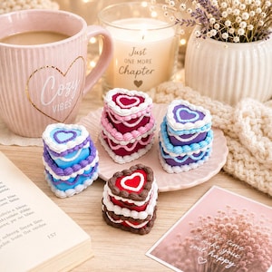 Op de afbeelding: Vier hartvormige doosjes met details die lijken op glazuur in rood, roze, blauw en paars, gerangschikt op een klein geschulpt bordje. De doosjes hebben gelaagde ontwerpen die doen denken aan miniatuurtaartjes met witte decoratieve randen.