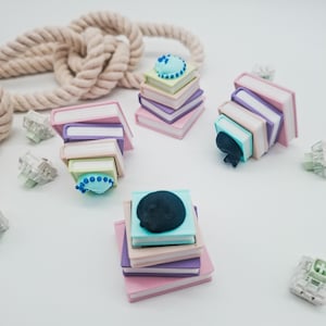 Op de afbeelding: Miniatuur stapels boeken in pastelkleuren, bekroond met kleine, decoratieve elementen. De boeken zijn roze, paars en lichtgroen, met een blauwe taart en een zwarte kat op sommige. Een touw is zichtbaar op de achtergrond.