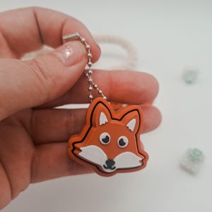 Könnte beinhalten: Ein fuchsförmiger Schlüsselanhänger. Der Anhänger ist orange mit weißen und schwarzen Akzenten und zeigt ein Fuchgesicht. Er hängt an einer silbernen Kugelkette. Der Hintergrund ist weiß.
