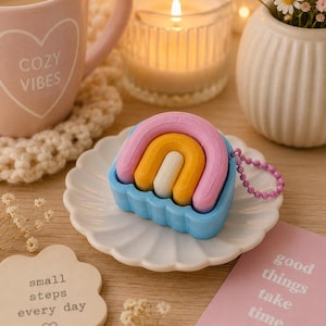 May include: A silicone protective case for wireless earbuds featuring a three-dimensional rainbow design with pink, yellow, and white arches set against a light blue base, complete with a small purple beaded chain attached to the side.