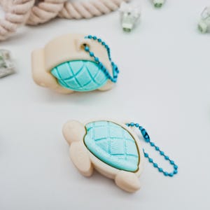 Könnte beinhalten: Zwei schildkrötenförmige Schlüsselanhänger. Die Schlüsselanhänger sind hellbeige mit einem türkisfarbenen Schalendesign. Jeder Schlüsselanhänger hat eine passende blaue Perlenkette. Der Hintergrund ist weiß mit einem Seil und anderen kleinen Objekten.
