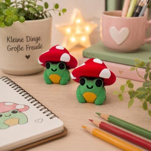 Könnte beinhalten: Zwei kleine grüne Froschfiguren mit gelben Bäuchen, die jeweils einen roten Fliegenpilzhut mit weißen Punkten tragen. Die Frösche haben schwarze Augen und einen lächelnden Mund. Sie stehen auf einer hellen Oberfläche neben einem Notizbuch.