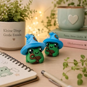 Könnte beinhalten: Zwei kleine grüne Froschfiguren mit leuchtend blauen Zauberhüten. Jeder Frosch hat große schwarze Augen, einen lächelnden Mund und hält einen braunen Stab. Die Figuren stehen auf einer Holzoberfläche und haben eine glatte Textur und runde Form.