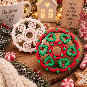 May include: Two circular mechanical gear fidget toys, one white with pink accents and one red with green accents, featuring a central ring surrounded by smaller interlocking gears with triangular cutouts and small pegs on the outer edges.