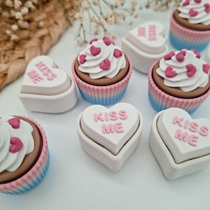 May include: A collection of Valentine's Day-themed treats. Cupcakes with white frosting and pink heart sprinkles sit next to heart-shaped boxes with the text "KISS ME" in pink. The cupcakes have blue and pink striped liners.