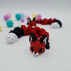 Könnte beinhalten: Ein rotes, schwarzes und weißes, bewegliches Fuchspielzeug mit detailliertem Design. Der Fuchs besteht aus mehreren verbundenen Segmenten, die eine flexible Bewegung ermöglichen. Bunte Pompons sind im Hintergrund verstreut.