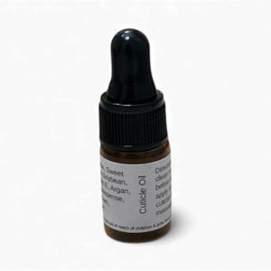 Op de afbeelding: Een klein bruin glazen flesje nagelriemolie met een zwarte druppelkap. Het etiket luidt "Cuticle Oil" en vermeldt de ingrediënten. Het flesje staat op een witte achtergrond.