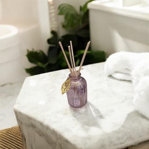 May include: A small, purple glass diffuser bottle with wooden reeds. A gold-colored leaf charm hangs from the bottle's neck. The diffuser sits on a white marble surface, with a white towel and greenery in the background.