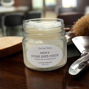 May include: A clear glass jar filled with sugar hand scrub. The jar has a white label with the text "MEN'S SUGAR HAND SCRUB" and "TEAKWOOD and MAHOGANY". The net weight is 8 oz.