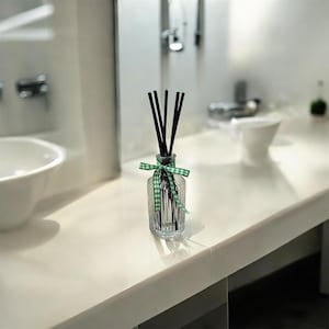 Może przedstawiać: Przezroczysta szklana butelka z czarnymi patyczkami i zielono-białą wstążką w kratkę. Butelka stoi na białym blacie w łazience. Szklana butelka ma pionowe linie wytrawione w szkle.