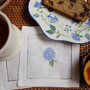 May include: Two white linen napkins with blue embroidered hydrangeas, a cup of tea, a plate of banana bread, and a bowl of orange slices on a woven wicker surface.