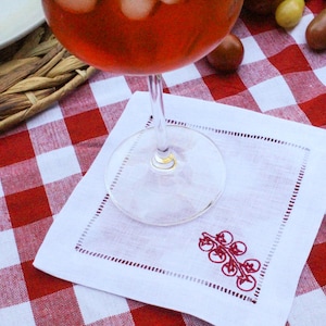 May include: A white linen cocktail napkin with a red embroidered tomato design, set on a red and white checkered tablecloth. A glass of red liquid with ice is above the napkin.