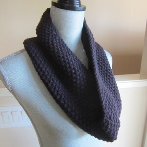 May include: A dark purple, hand-knitted infinity scarf displayed on a mannequin. The scarf is made of thick yarn, with a visible knit texture. The color is a deep, rich shade of purple, suitable for cold weather.