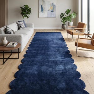 Puede incluir: Una alfombra larga de color azul marino con un borde festoneado. La alfombra está colocada sobre un suelo de madera clara en un salón. La habitación también cuenta con un sofá gris, sillas y plantas.