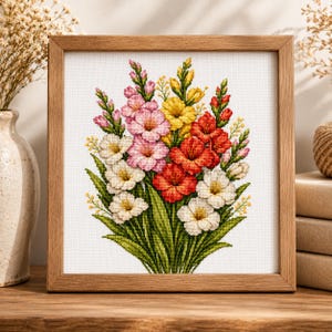 May include: A square wooden frame containing a cross-stitch embroidery of a vibrant floral bouquet. The design features gladiolus flowers in shades of pink, yellow, red, and white, accented with green leaves and stems on a white fabric background.