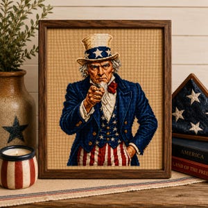 May include: A framed cross-stitch embroidery depicting Uncle Sam wearing a blue coat, a red and white striped vest, and a top hat with stars. The figure is pointing forward with a serious expression, rendered in detailed thread work on a tan fabric.