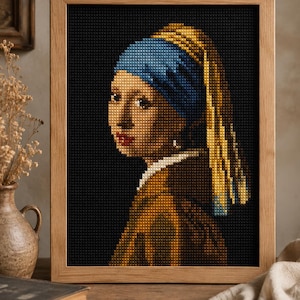 Op de afbeelding: Een ingelijst kruissteek borduurwerk met een portret van een persoon met een blauwe hoofddoek en een geel hoofddeksel tegen een effen zwarte achtergrond. Het kunstwerk zit in een lichtgekleurde houten lijst met een zichtbare rasterstructuur.