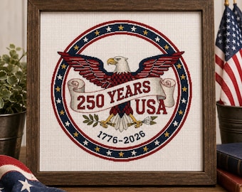 May include: A framed cross-stitch embroidery featuring a bald eagle with spread wings, clutching an olive branch and arrows. A banner across the eagle reads 250 YEARS USA, with 1776-2026 below, all enclosed in a circular border of blue, red, and stars.