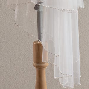 May include: A white, sheer, multi-layered tulle wedding veil featuring a delicate scalloped edge adorned with small, evenly spaced white pearls. The veil is draped over a stand, showcasing the fine mesh fabric and the pearl-embellished trim.