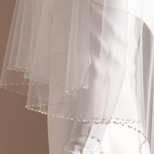 Puede incluir: Un velo de novia blanco y transparente, con varias capas. Los bordes del velo están adornados con pequeñas y delicadas perlas. El velo está drapeado sobre una prenda blanca, creando un efecto suave y fluido. La estética general es elegante y clásica.