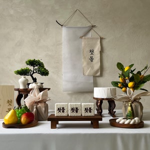 May include: A traditional Korean offering table with a white cake, fruit, and rice cakes. The table is decorated with a bonsai tree, a vase, and a banner with the Korean text "축복" which means "blessings".