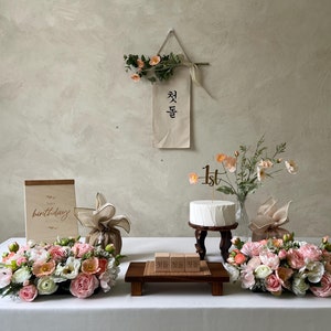 May include: A white cake with a wooden "1st" topper sits on a wooden stand. The cake is decorated with pink and peach flowers. A wooden sign with the text "Happy birthday" is on the table. There are also two bouquets of pink and peach flowers, a wooden sign with Korean text, and a brown gift bag with a white ribbon.